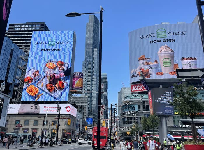 Billboards in a bustling urban area announce the opening of Shake Shack, displaying images of burgers, fries, and milkshakes