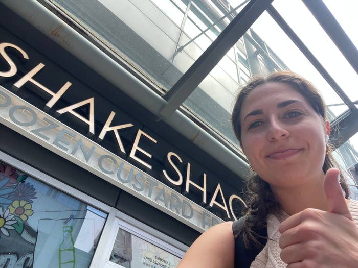 A woman smiles and gives a thumbs up in front of a Shake Shack sign at an outdoor location