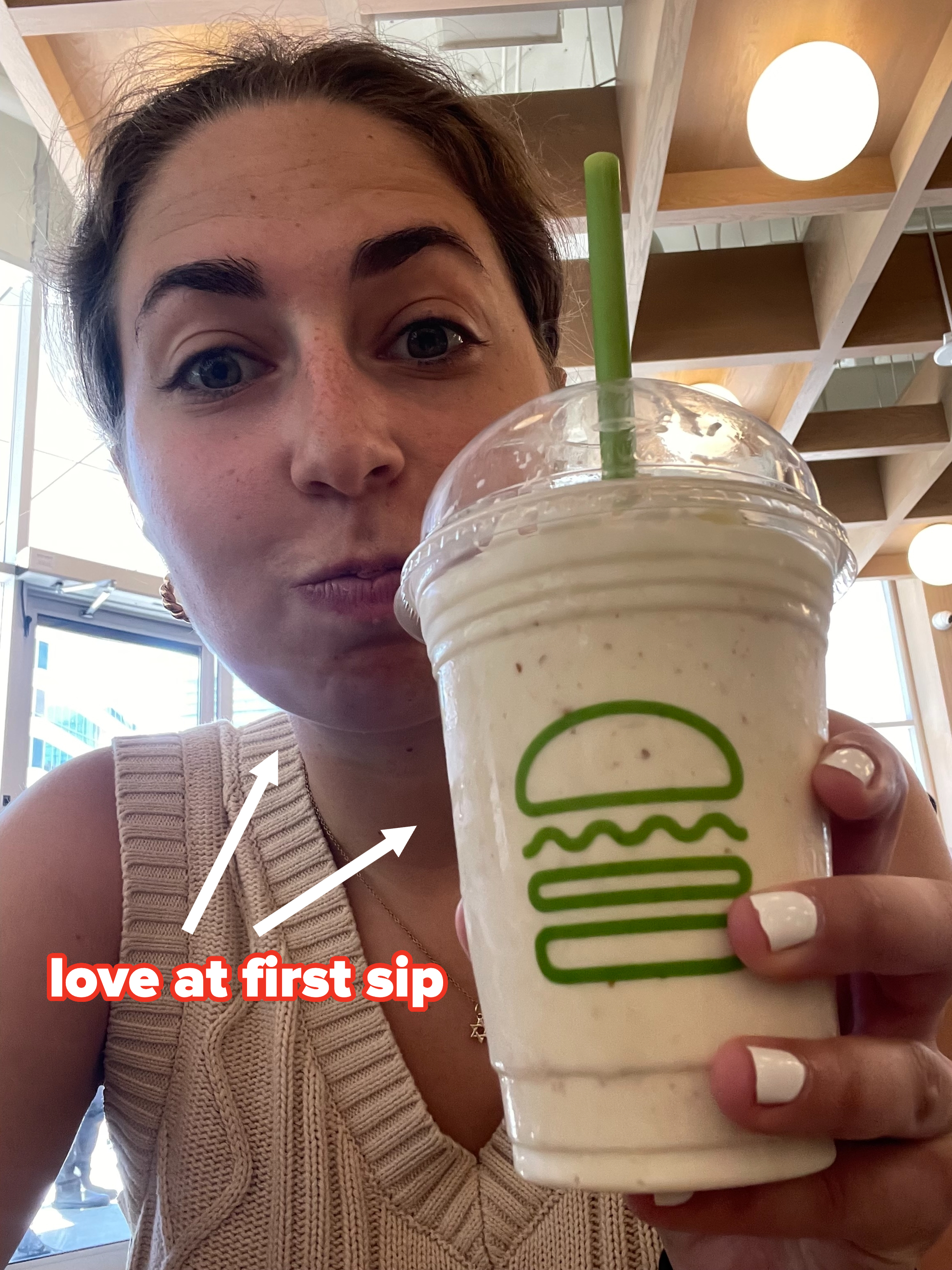 Person holding a milkshake with a green straw in a cup featuring the Shake Shack logo, smiling into the camera indoors