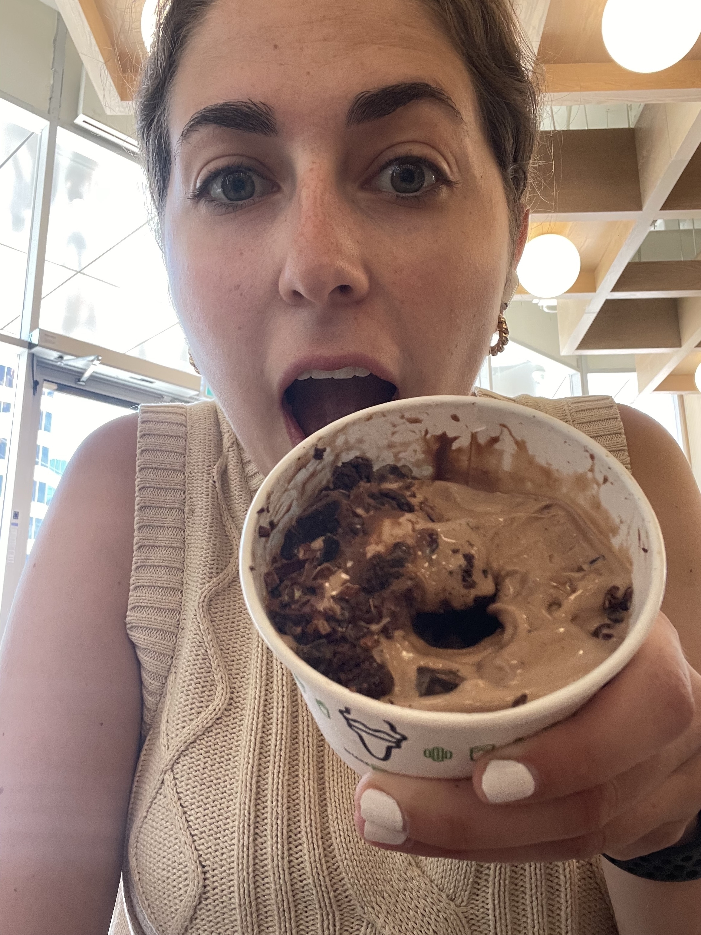 A woman in a knitted top holds a cup of chocolate ice cream with chocolate toppings, looking excited to take a bite