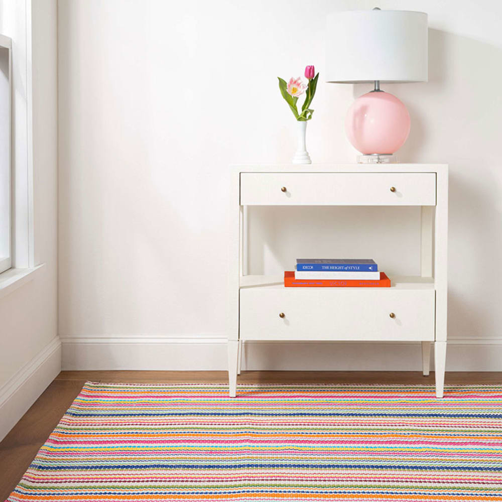 A modern nightstand with a pink lamp, a small vase with flowers, and two books on one shelf. A striped rug lies on the floor in front