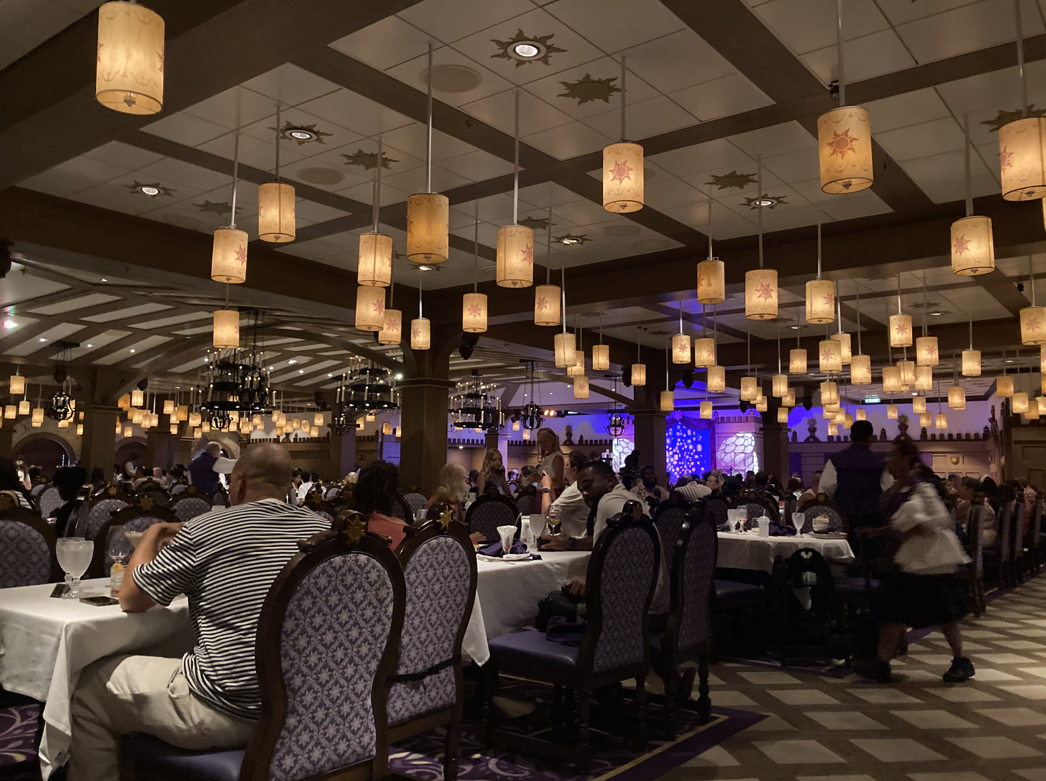 A busy restaurant dining area with numerous hanging lanterns, many patrons seated at tables, and a few people walking around