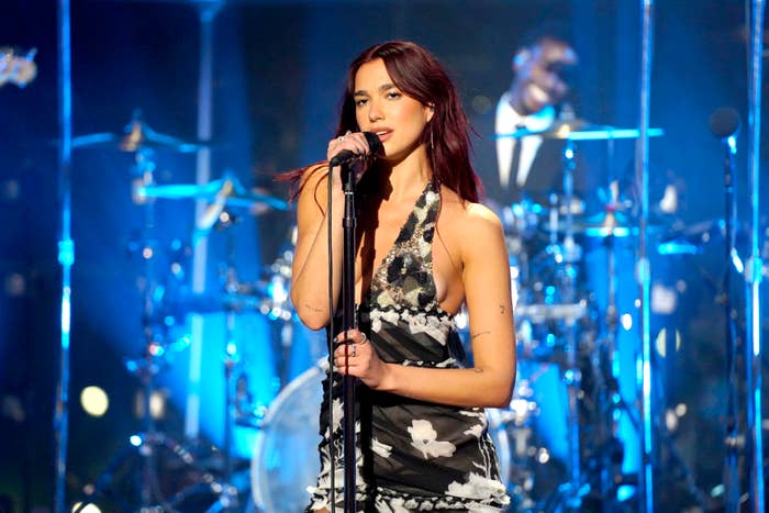 Dua Lipa performs on stage, wearing a stylish halter dress with floral patterns. A drummer is visible in the background