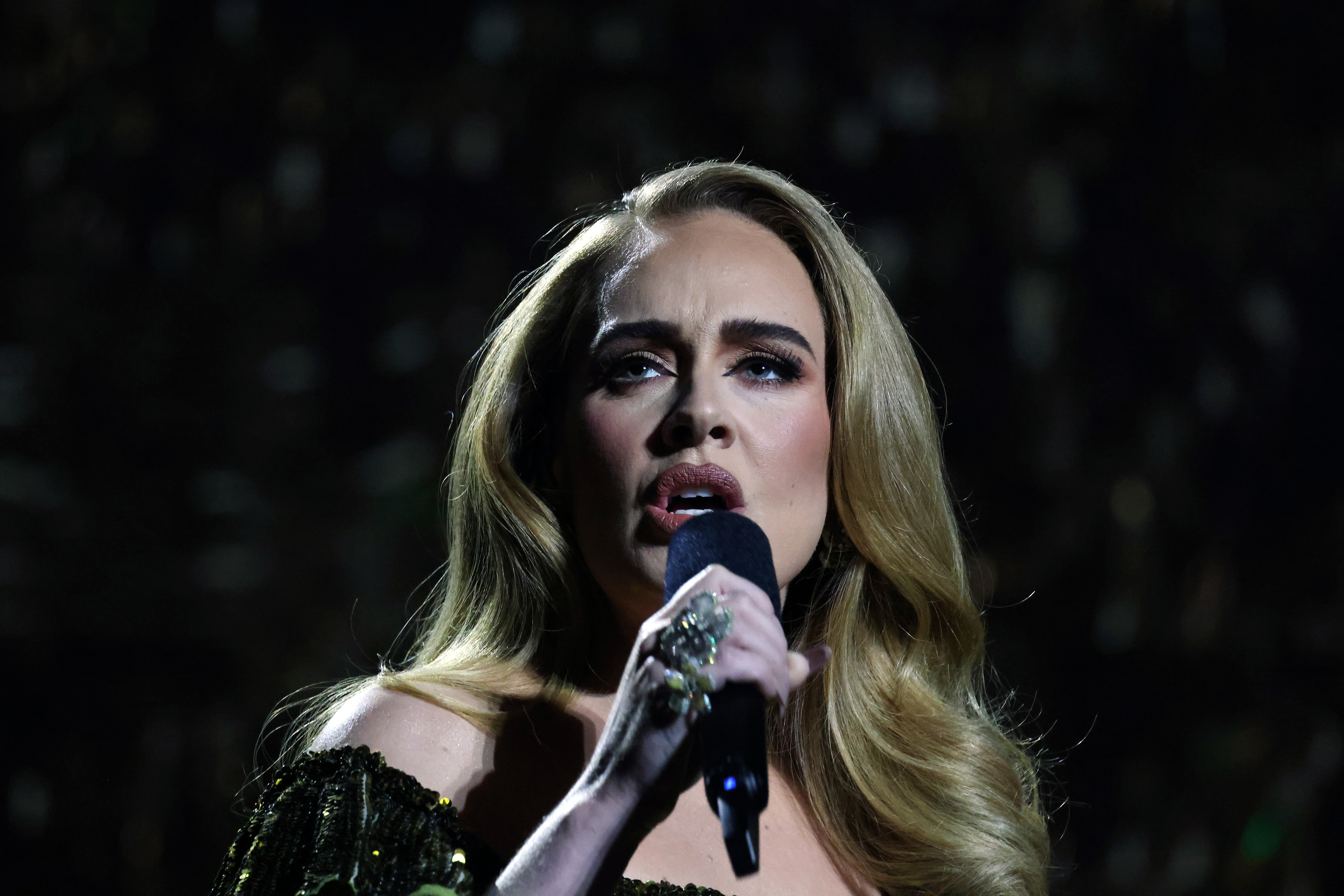 Adele sings passionately into a microphone, wearing an elegant off-the-shoulder evening gown