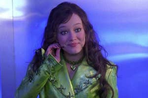 Hilary Duff smiles while wearing a metallic, futuristic green outfit with a heart necklace and holding a small device to her ear