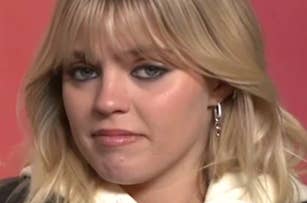 A woman with shoulder-length blonde hair and bangs looks directly at the camera, wearing a hoodie and hoop earrings against a red background