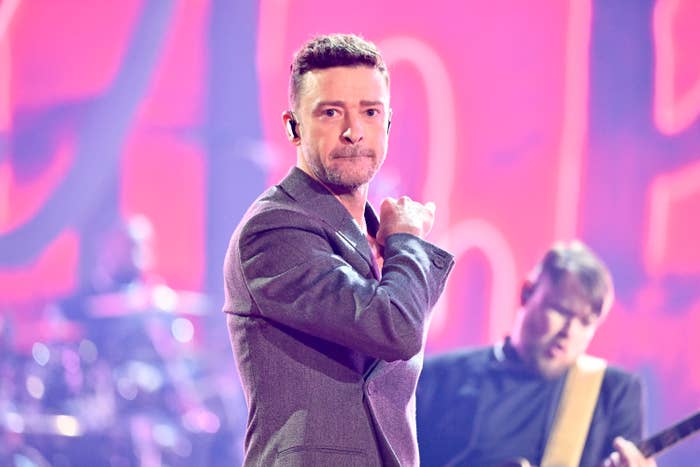 Justin Timberlake performs on stage, wearing a suit jacket. A guitarist plays in the background