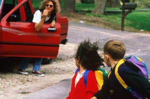 An adult in sunglasses leans out of a red car talking to two children with backpacks, one in a red blazer and the other in a black sweatshirt, walking hand in hand