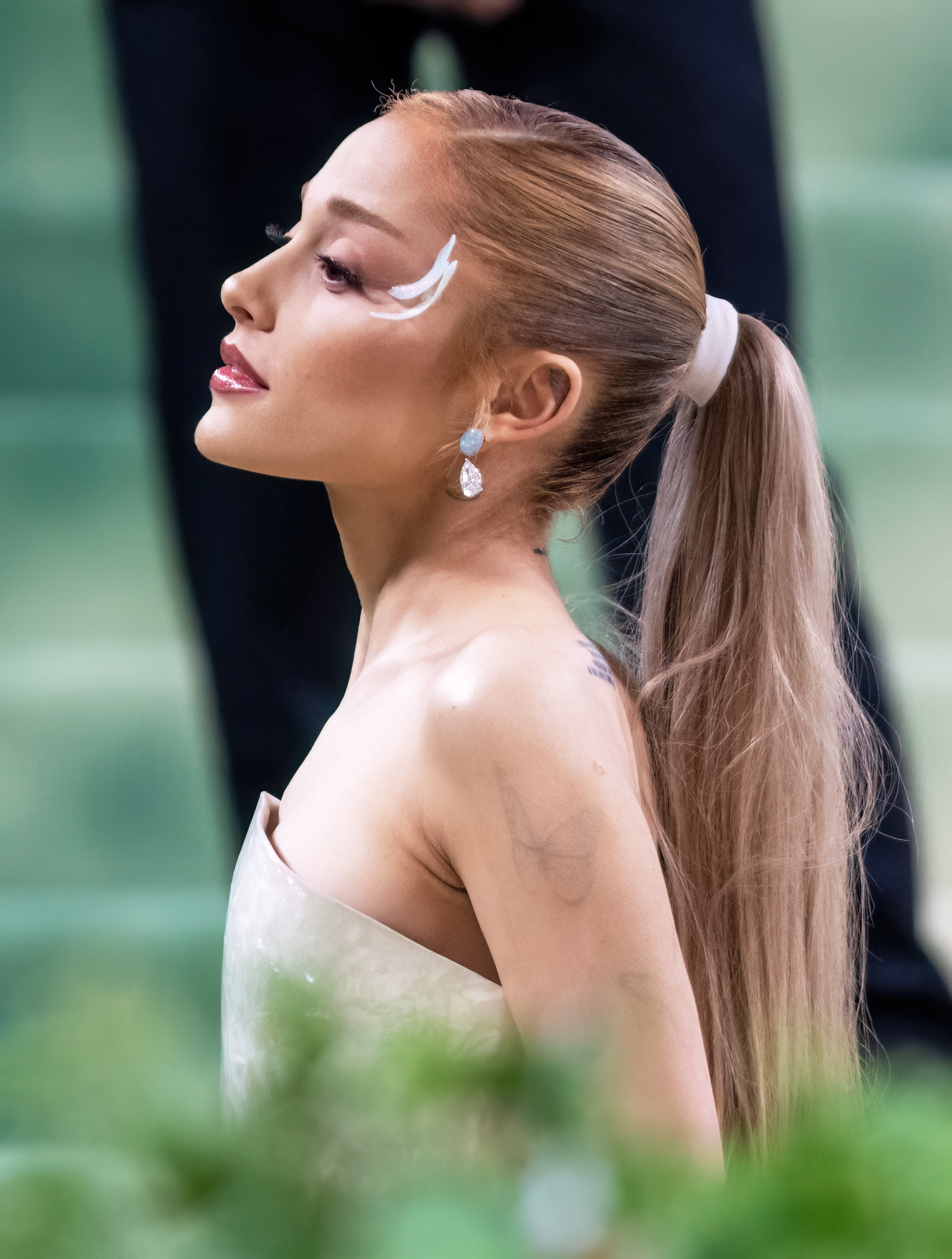 Ariana Grande at a public event; she wears an off-shoulder dress, long ponytail, and bright makeup with a bold winged eyeliner
