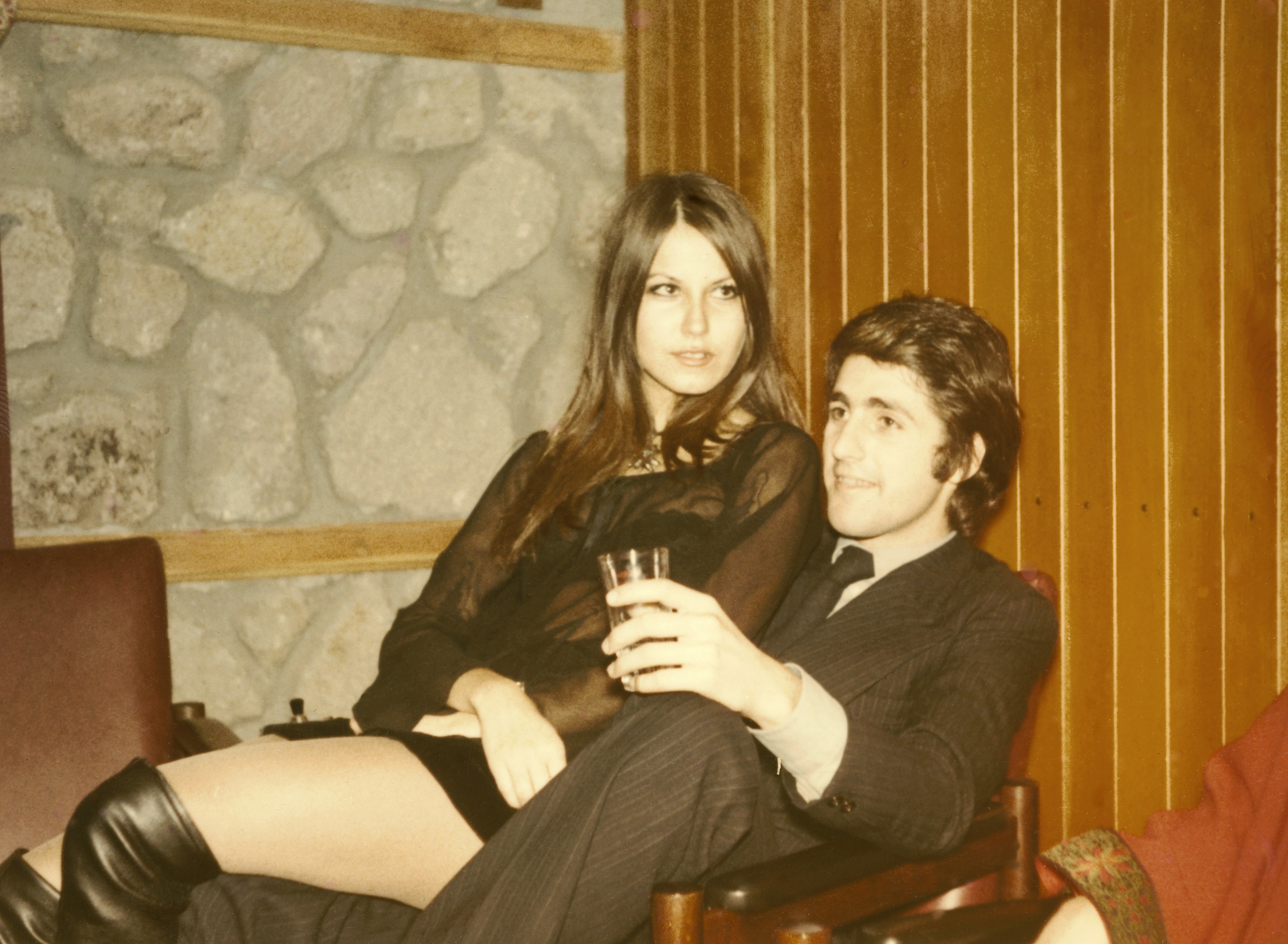 A woman in a short dress and knee-high boots sits on a man&#x27;s lap. Both are holding drinks and looking off-camera