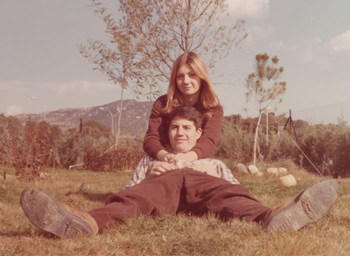 A couple in a grassy field; a woman sits behind a man, both smiling and embracing each other. The mountains are visible in the background