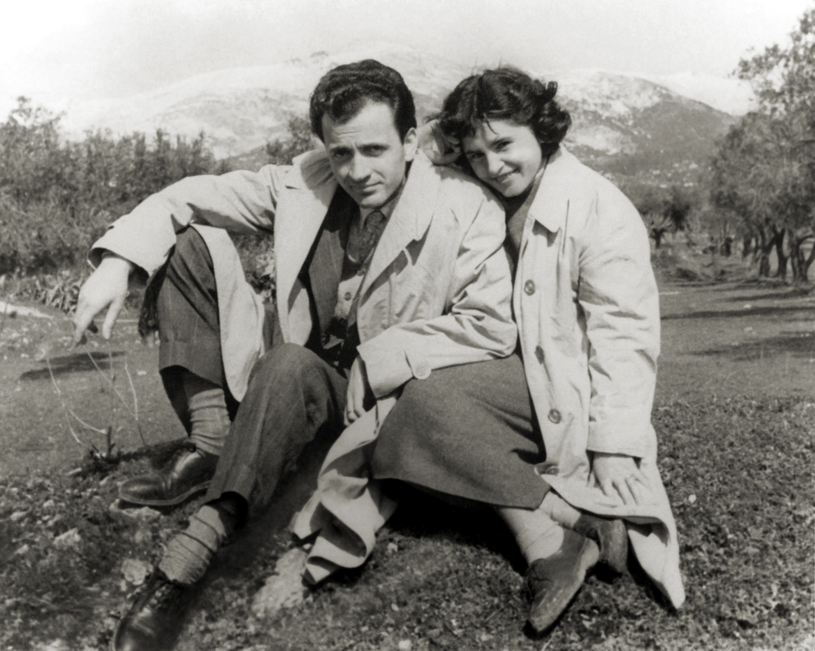 A man and woman, both wearing trench coats, sit closely together on the ground outdoors, smiling