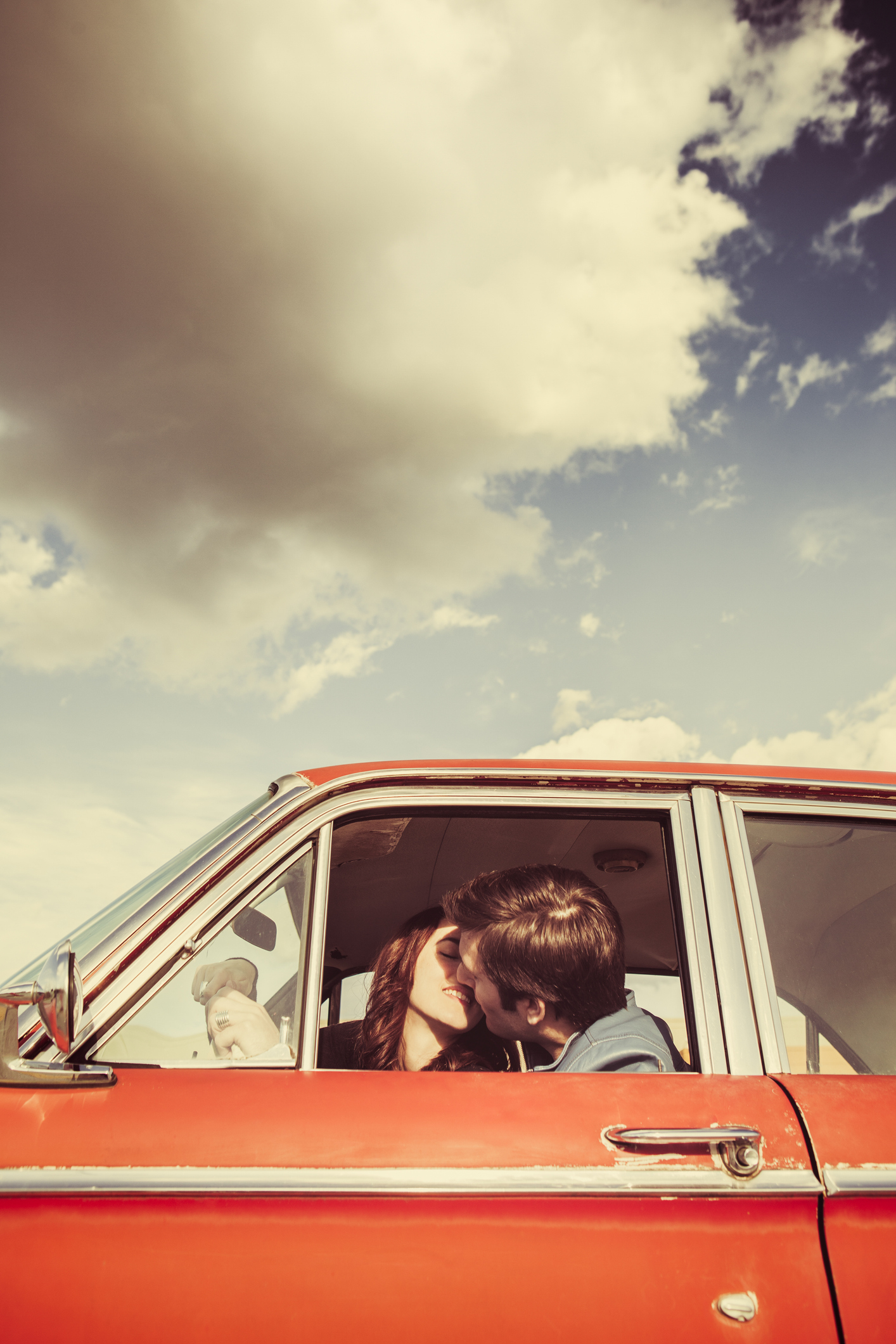 A couple kisses inside a parked vintage car with the window down under a partly cloudy sky