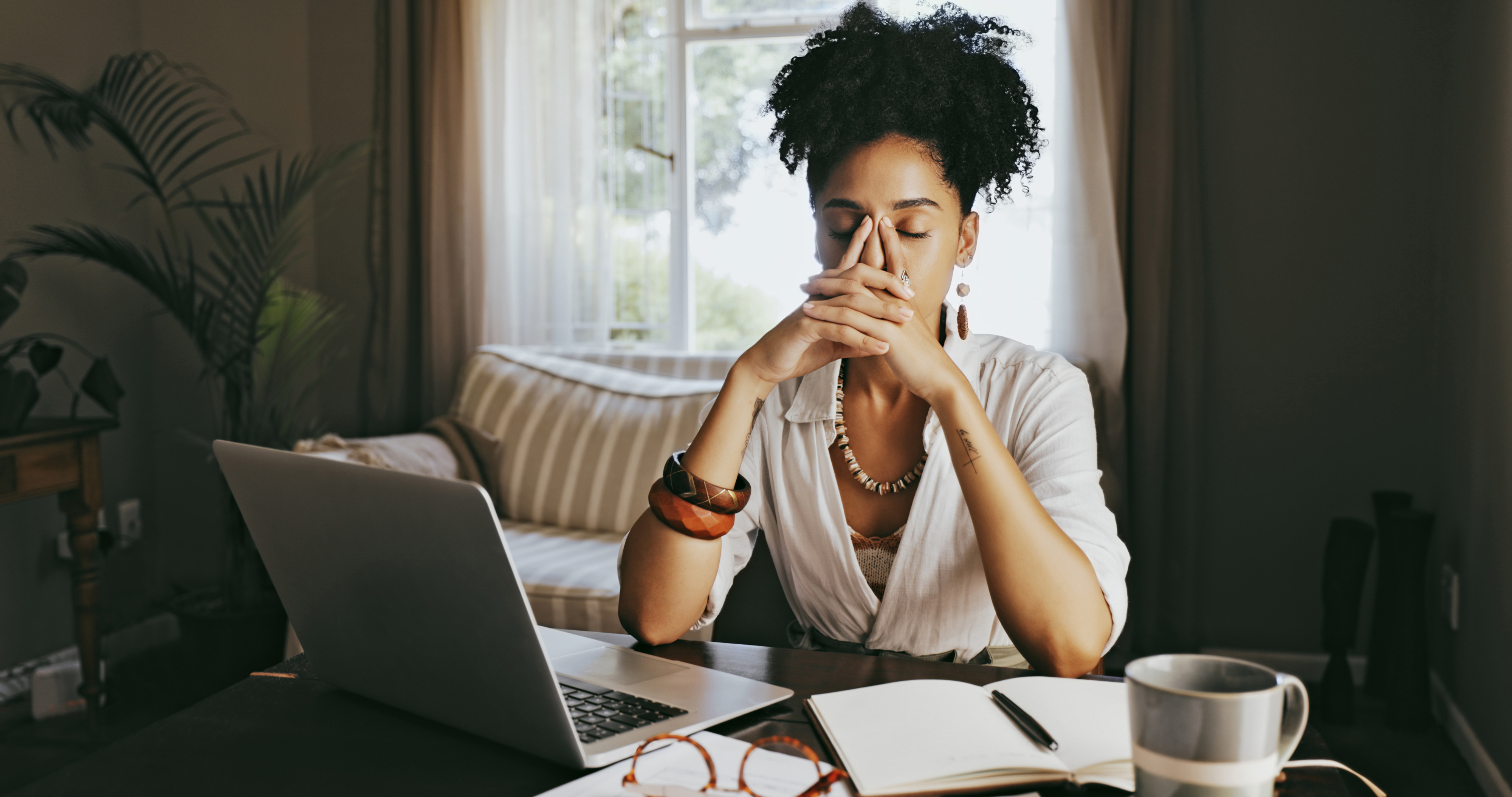 A woman sitting at a desk with a laptop, notebook, and coffee, appears deep in thought with hands clasped in front of her face