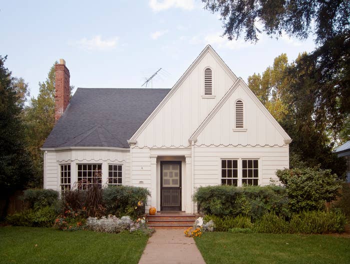 Small white house with steep roof, chimney, and well-kept front yard with bushes and flowers. A pumpkin sits on the front steps beside the door