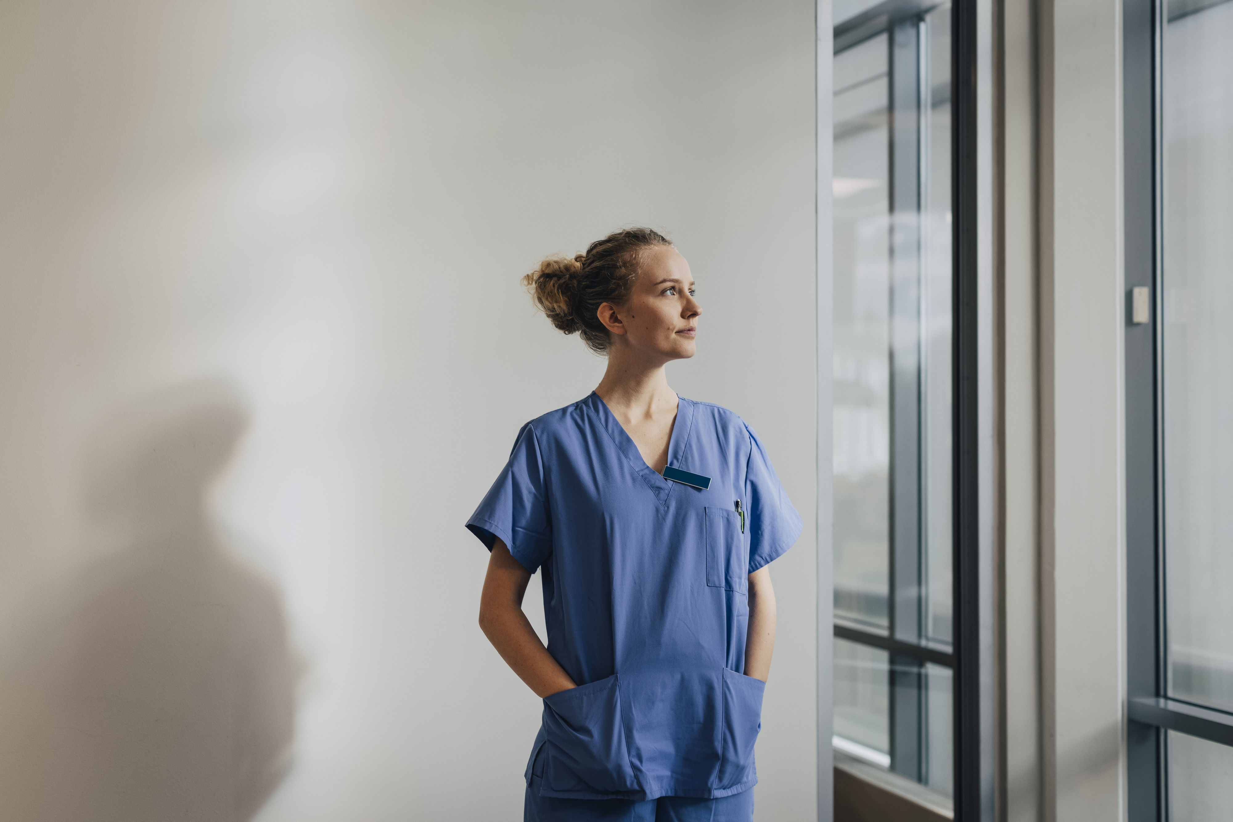 A person in medical scrubs stands by a window in a hospital or clinic, looking thoughtfully to the side