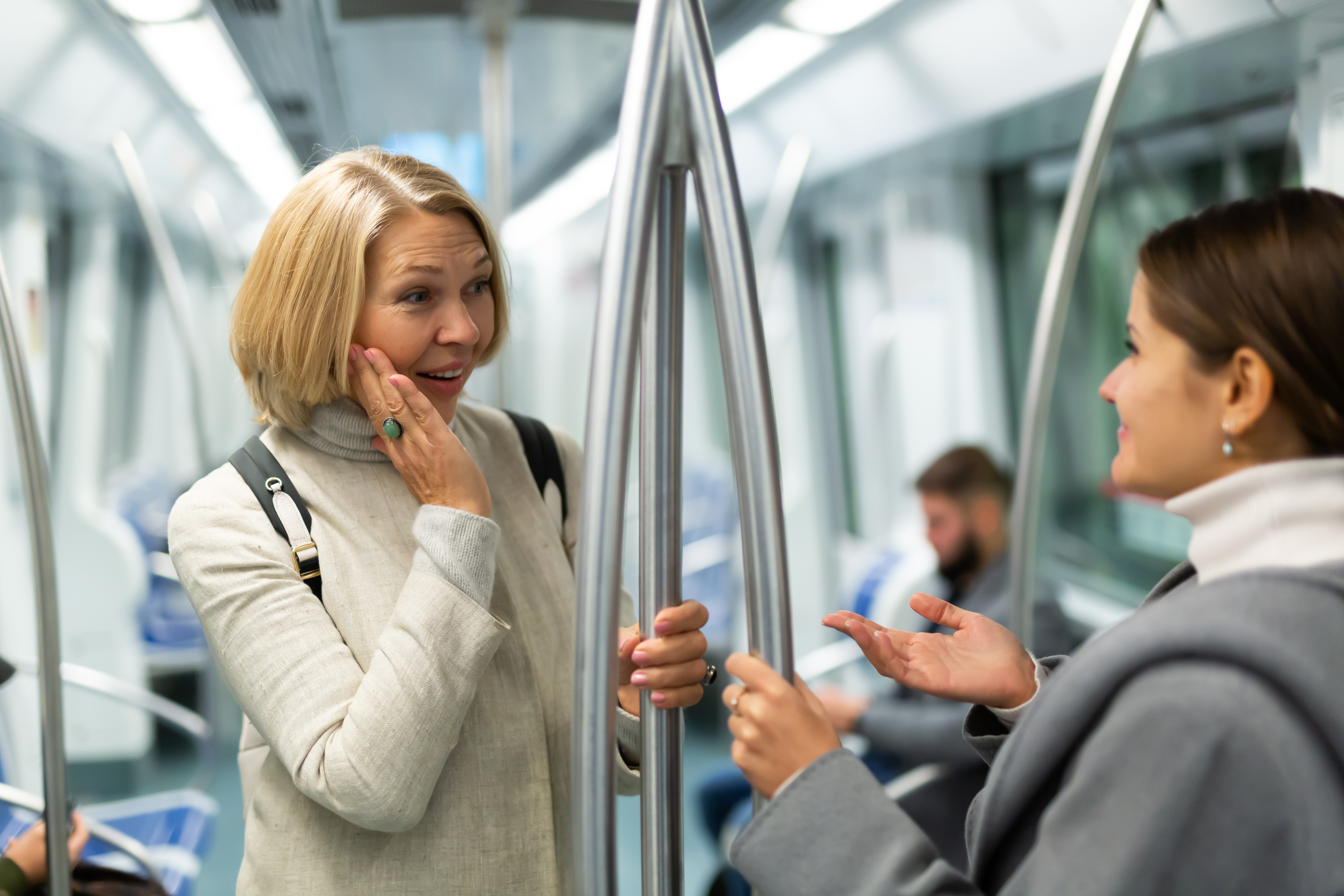 Two women are conversing and smiling while standing inside a subway train, both holding onto poles for support. Other passengers are seated in the background