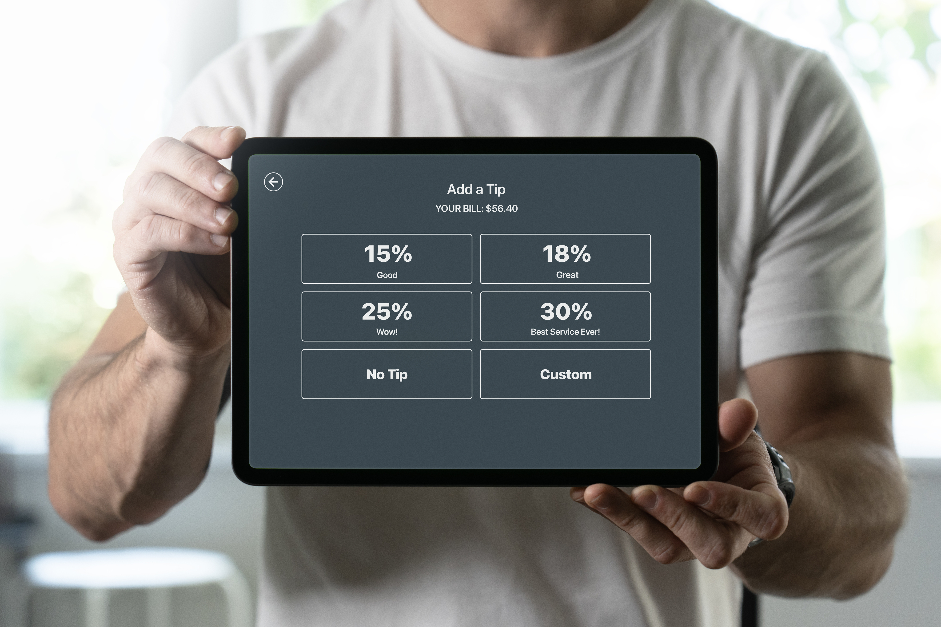 A person holds a tablet displaying a tip selection screen for a $56.40 bill, with options for 15%, 18%, 25%, 30%, no tip, and custom tip amounts