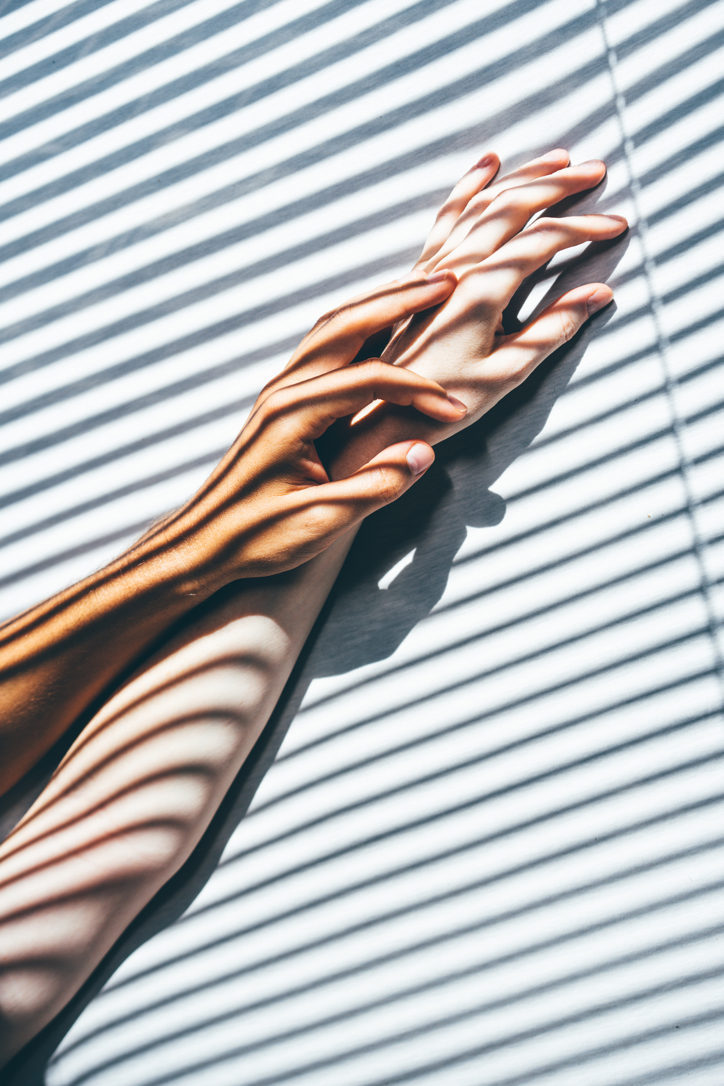 Two interlaced hands with light and shadow effects from blinds, creating an artistic pattern of stripes across the skin