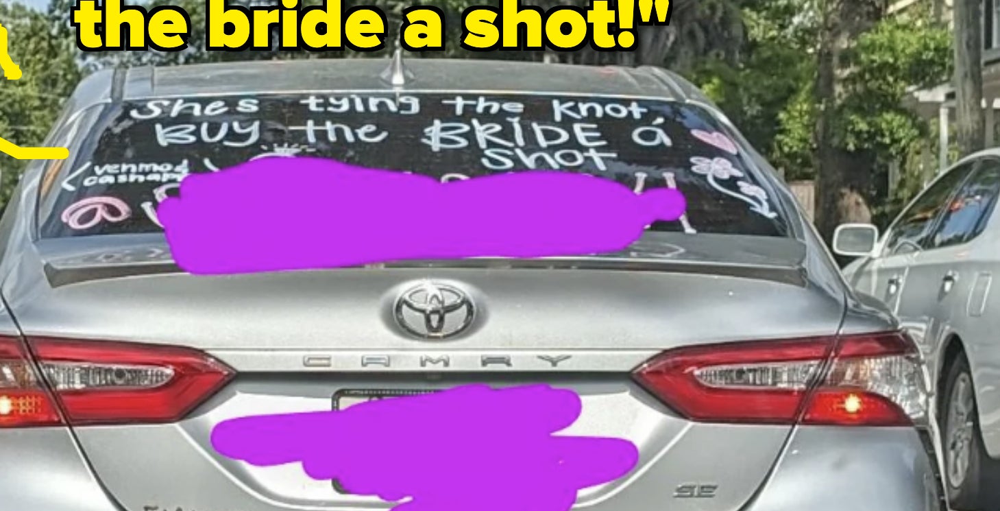 Car with &quot;She&#x27;s tying the knot! Buy the bride a shot!&quot; written on rear window driving on street. Trees, other cars, and traffic lights are visible