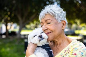 An elderly person happily hugs a small white dog outside in a park