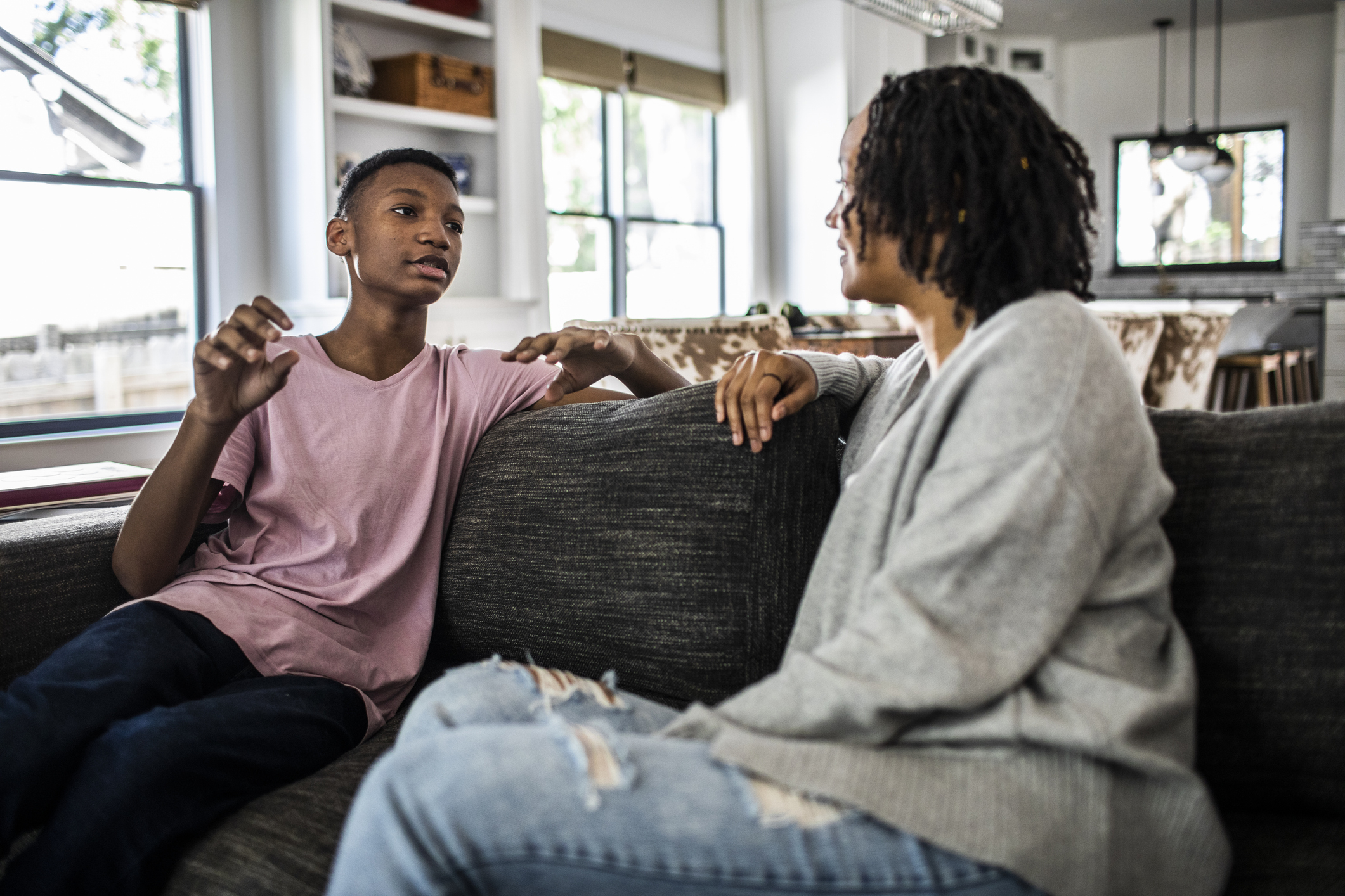 A teenager and an adult are sitting on a couch in a casual home setting, engaged in a conversation. Names are unknown