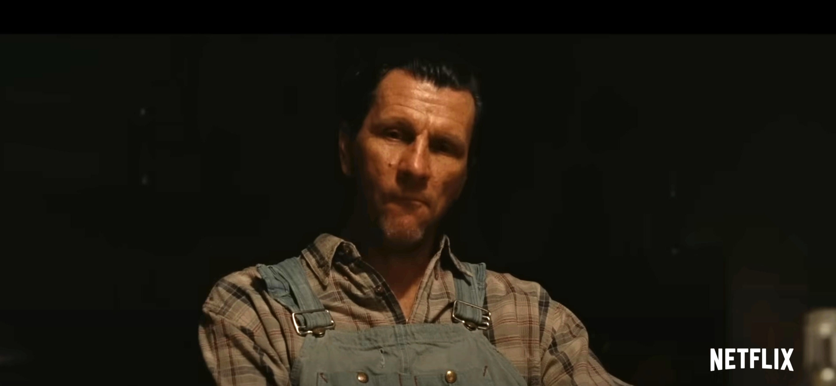 A man wearing overalls and a plaid shirt in a scene from a Netflix production. The scene is dimly lit