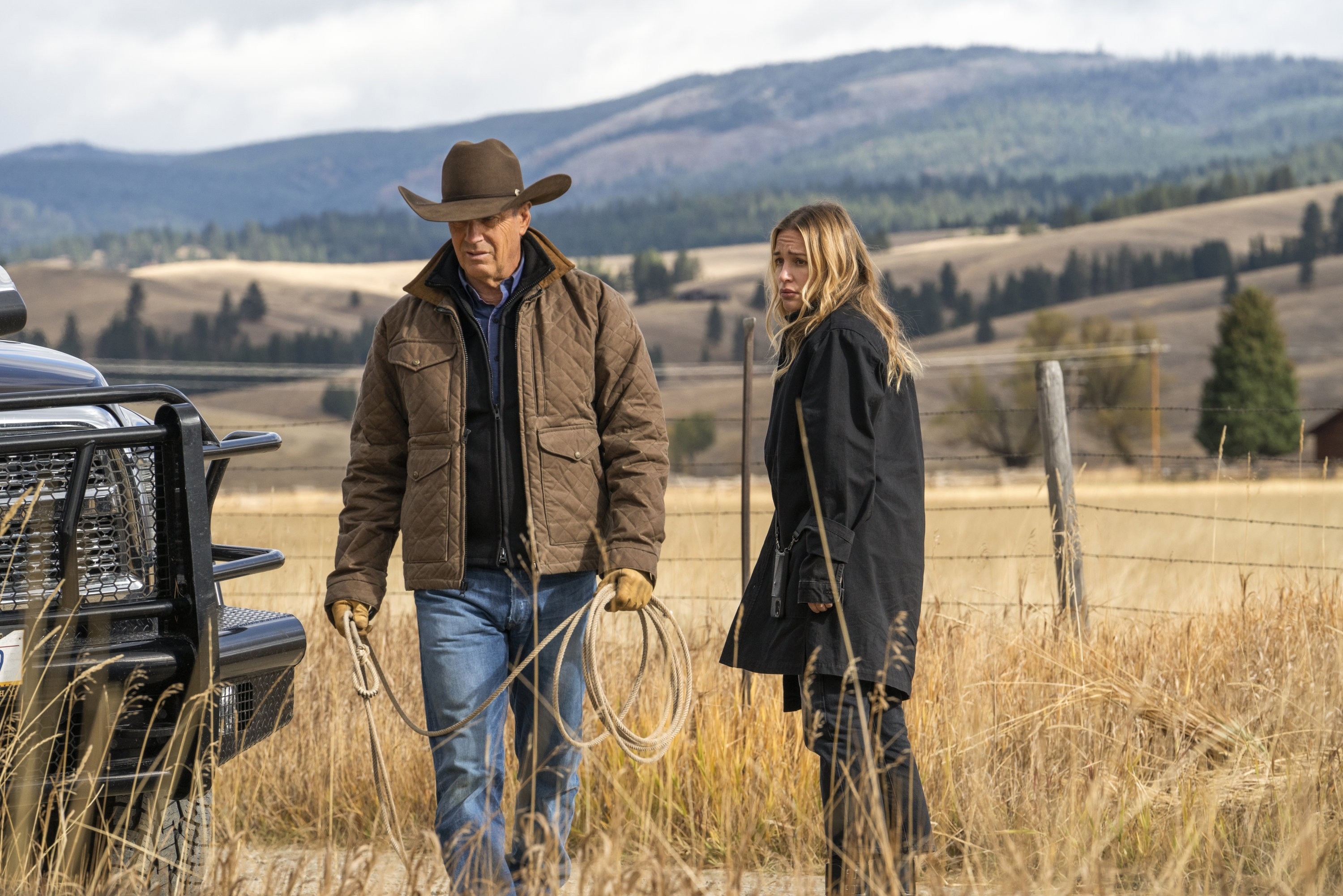 Kevin Costner and Piper Perabo standing outdoors in a rural setting with mountains in the background while filming. Costner holds a rope, in a cowboy hat and jacket