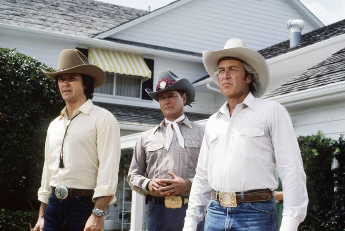 Three men in cowboy hats and western attire stand in front of a house