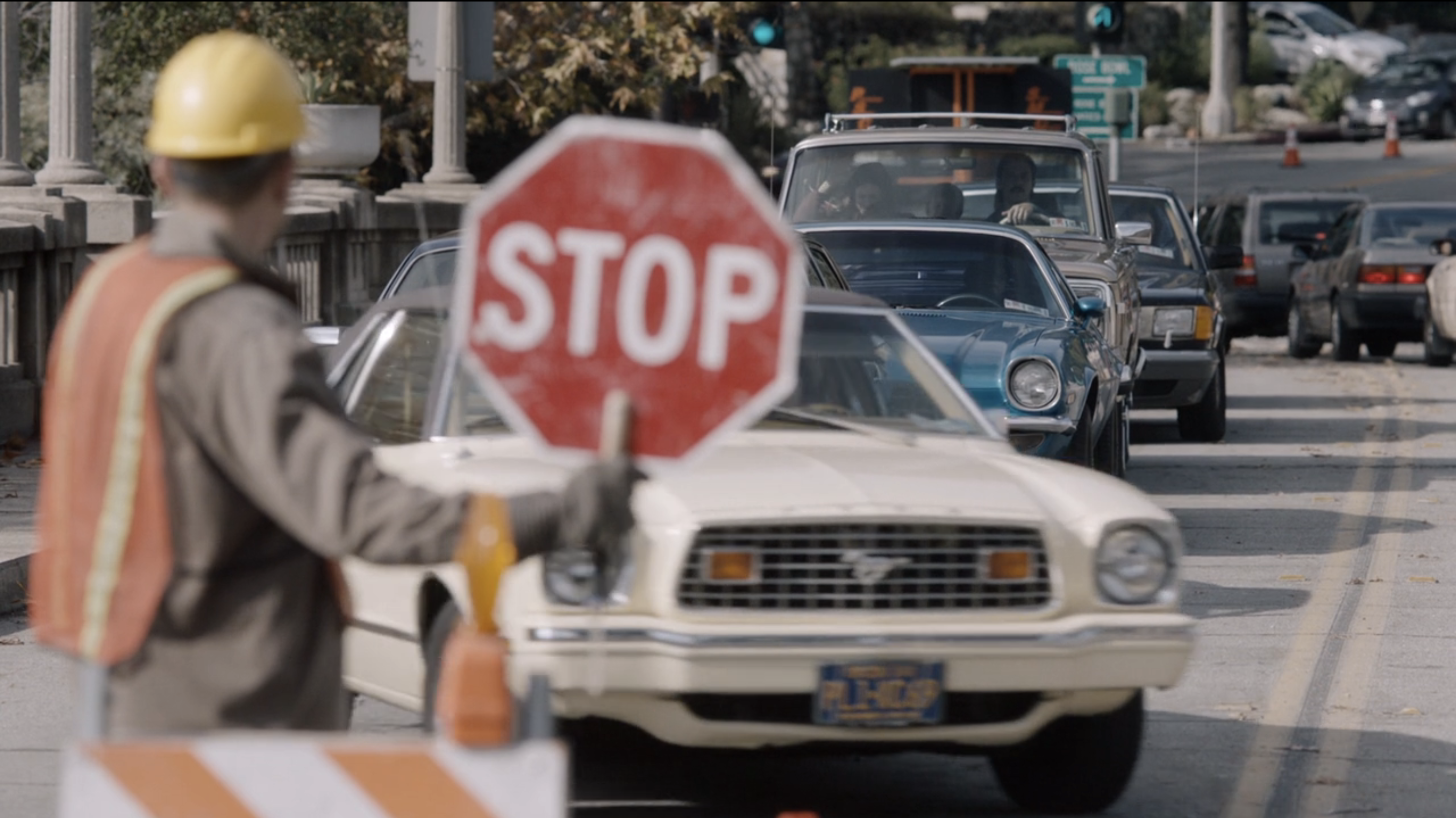A construction worker holds a &quot;STOP&quot; sign, halting a line of cars on a city street, including a classic Ford Mustang with license plate at the front