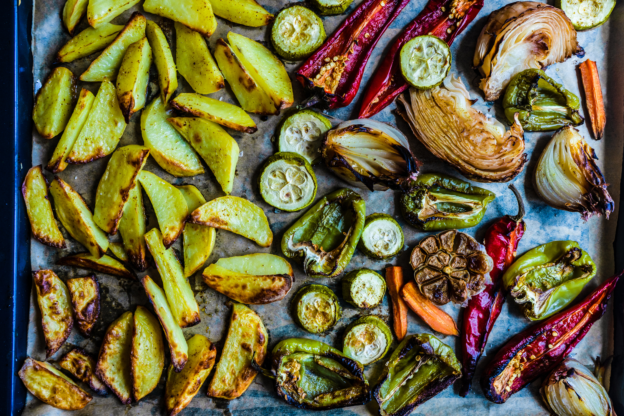 Roasted vegetables including potato wedges, zucchini slices, bell pepper halves, onion wedges, red and green chilies, garlic bulb, and carrots on a baking sheet