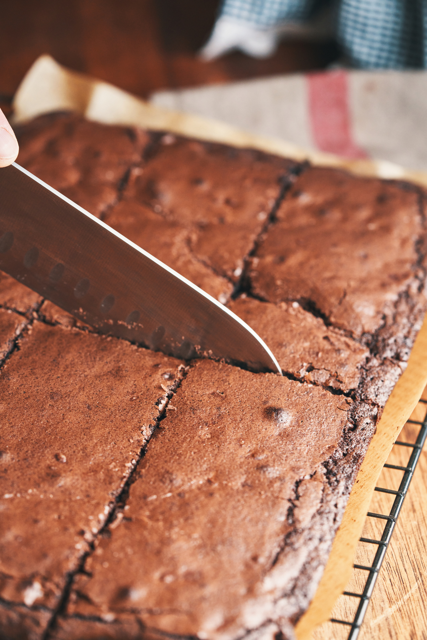 A knife is cutting a freshly baked tray of brownies into squares on a cooling rack