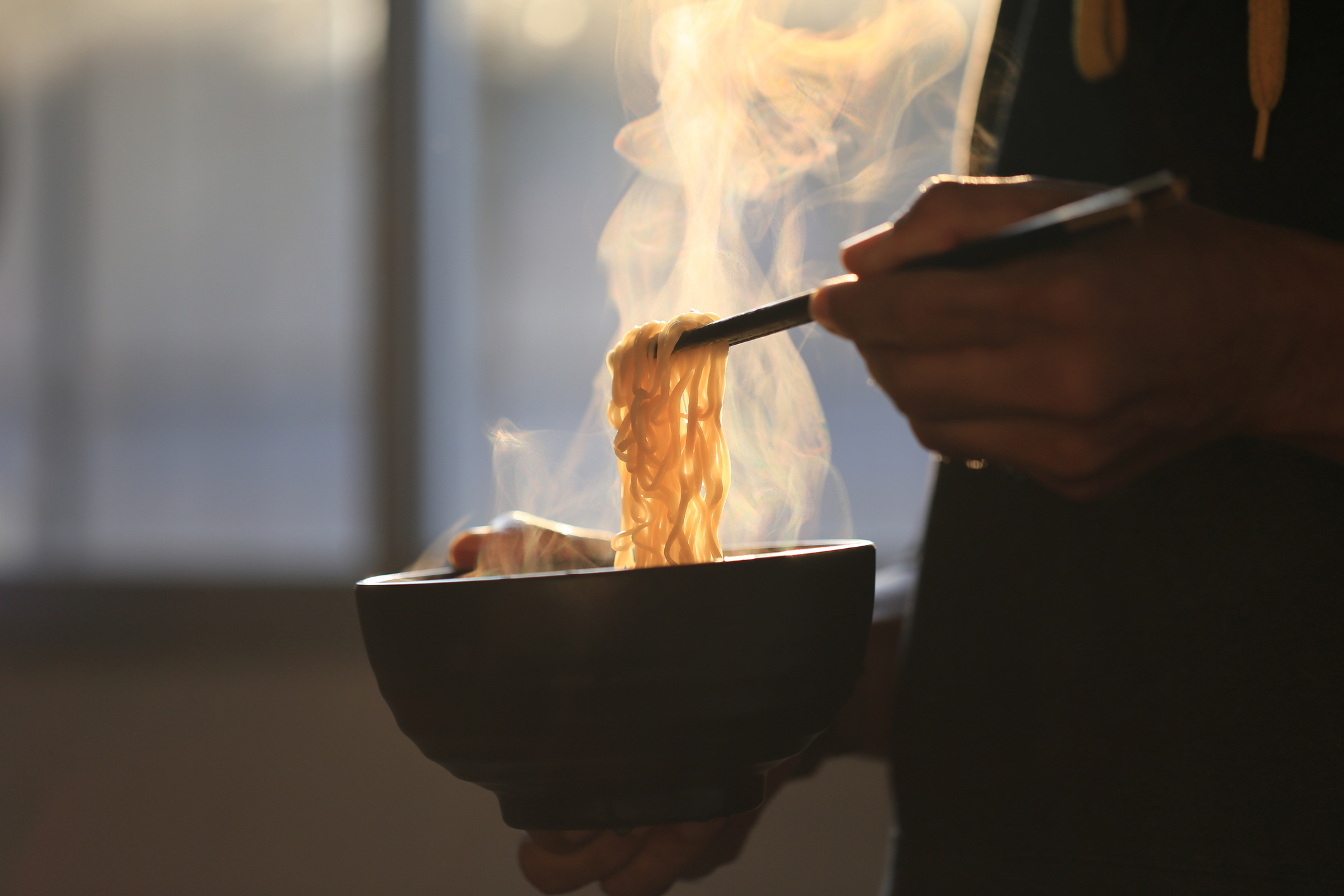 A person holding a bowl of steaming noodles with chopsticks by a window
