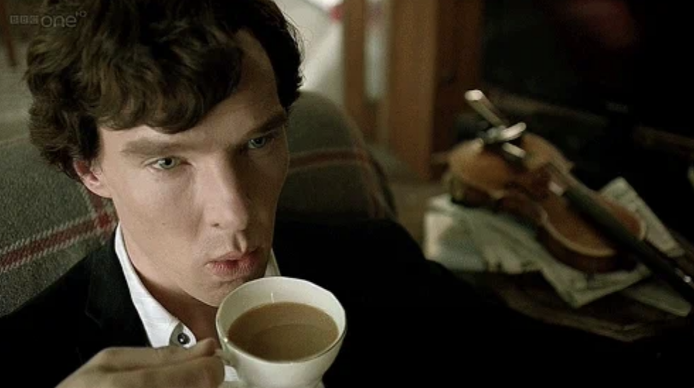 Benedict Cumberbatch, as Sherlock Holmes, sits on a couch, holding a cup of tea, with a violin and papers on a table behind him