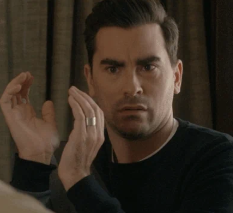 Dan Levy, dressed casually, looks shocked with his hands raised