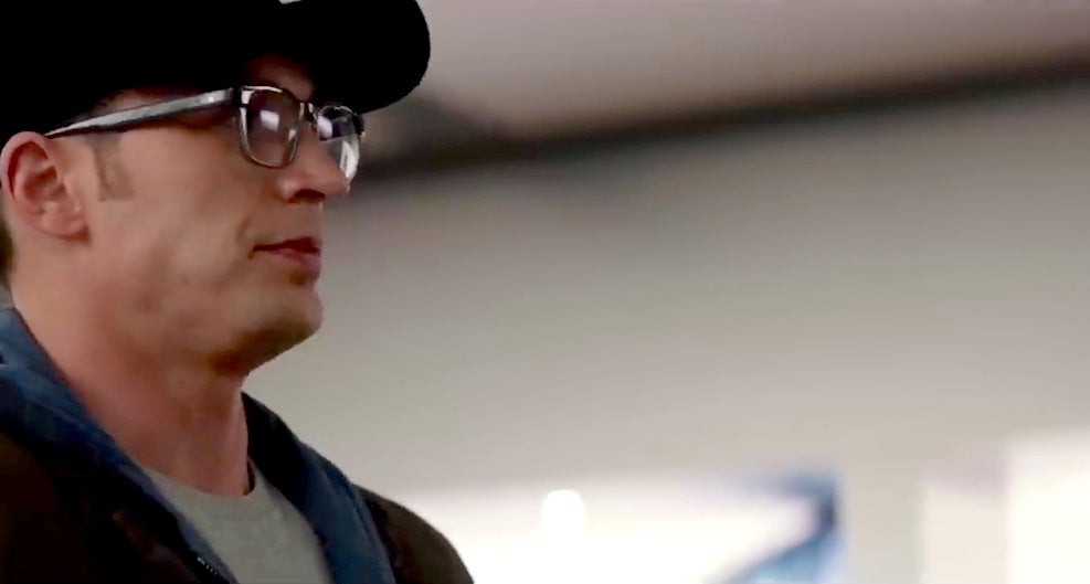 Man with glasses and cap, wearing a casual hoodie and jacket, looks to his right in an indoor setting
