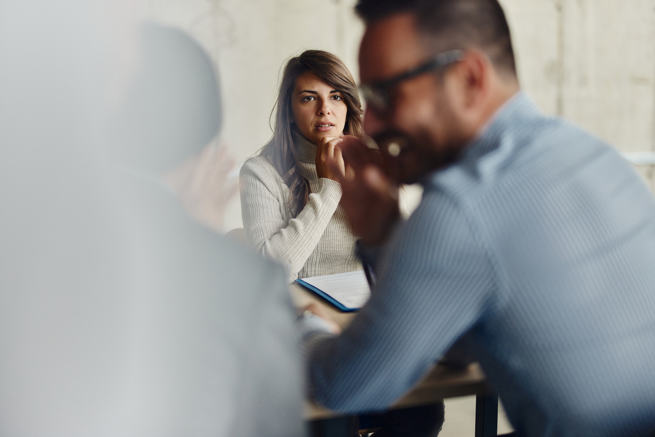 A woman sits thoughtfully at a table, looking at a man in a discussion setting. Office environment suggests a business meeting or brainstorming session