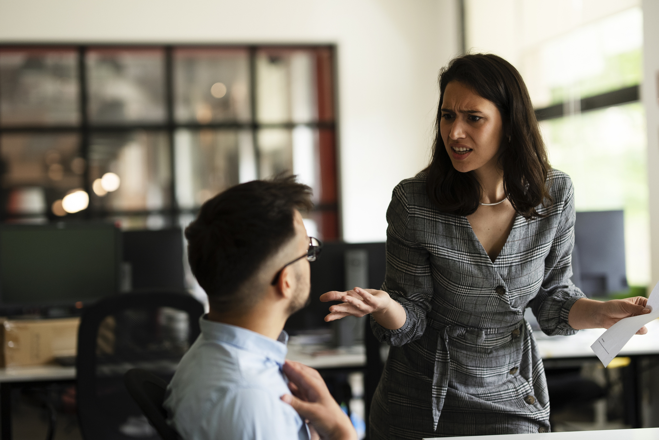 A woman in office attire holds a paper and gestures, appearing frustrated, while speaking to a seated man in glasses, in a modern office setting