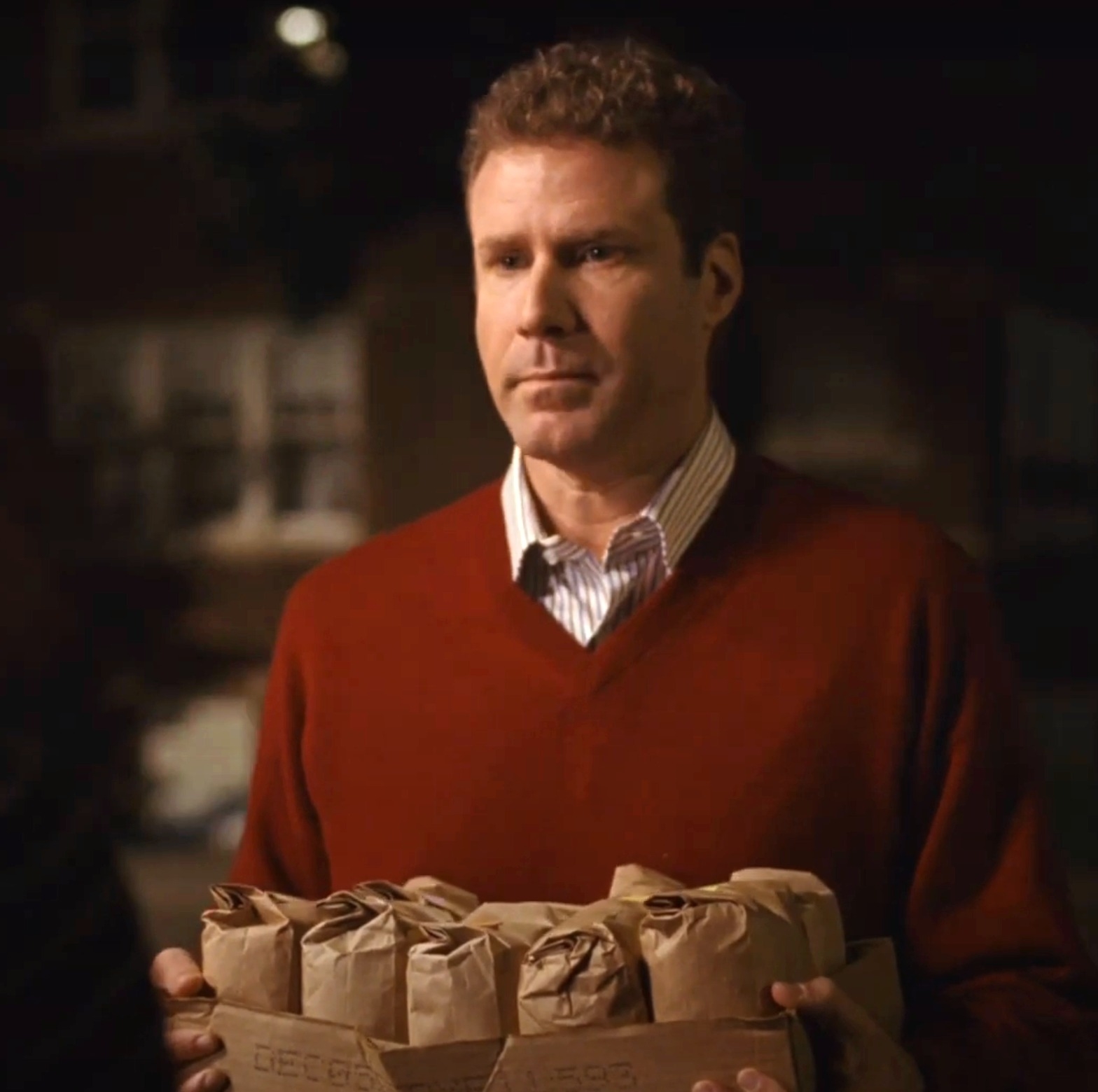 Will Ferrell holding a cardboard 6-pack carrier with paper-wrapped items