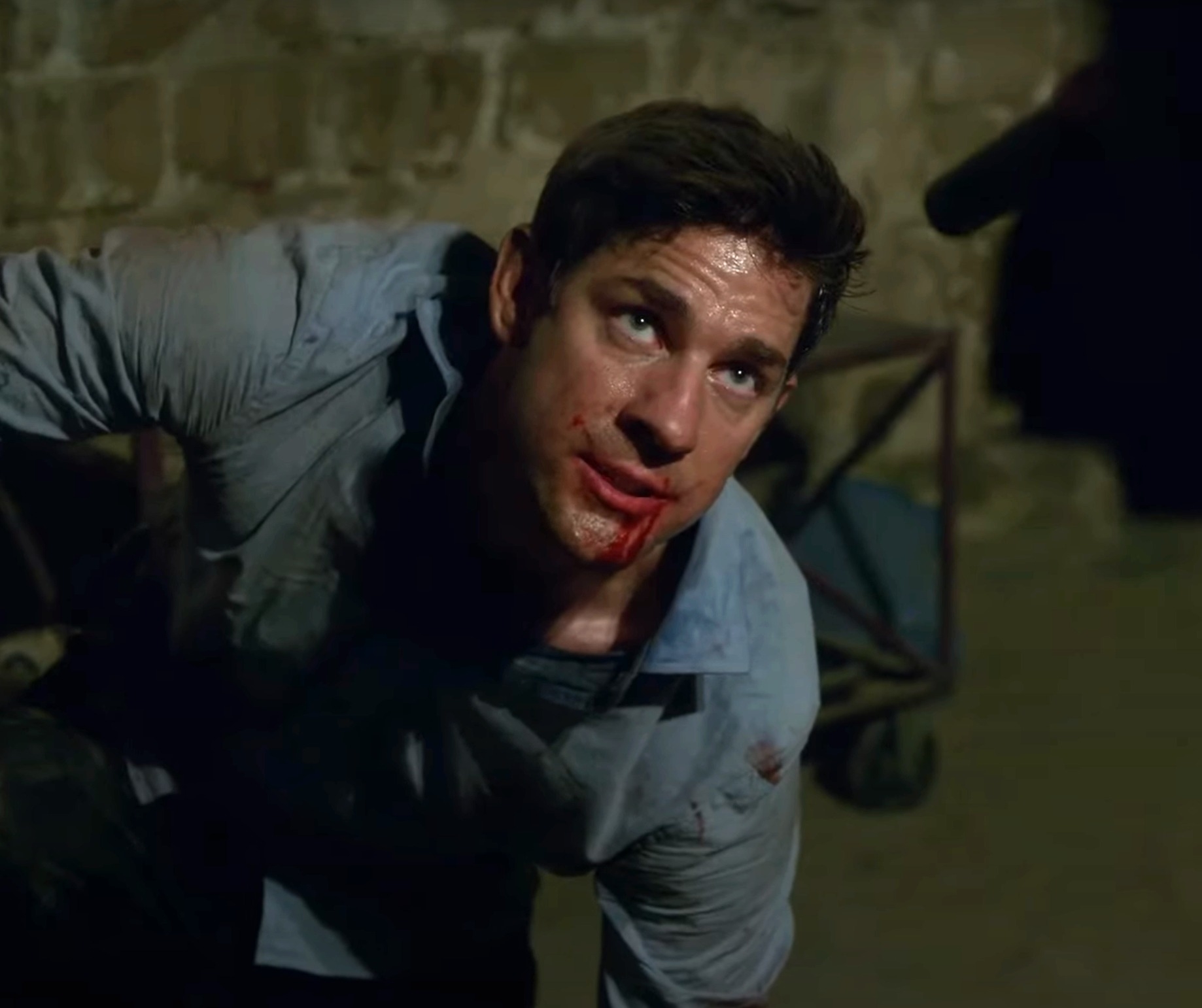 John Krasinski, disheveled and injured, is on his knees with blood on his face, looking up in a tense moment.
