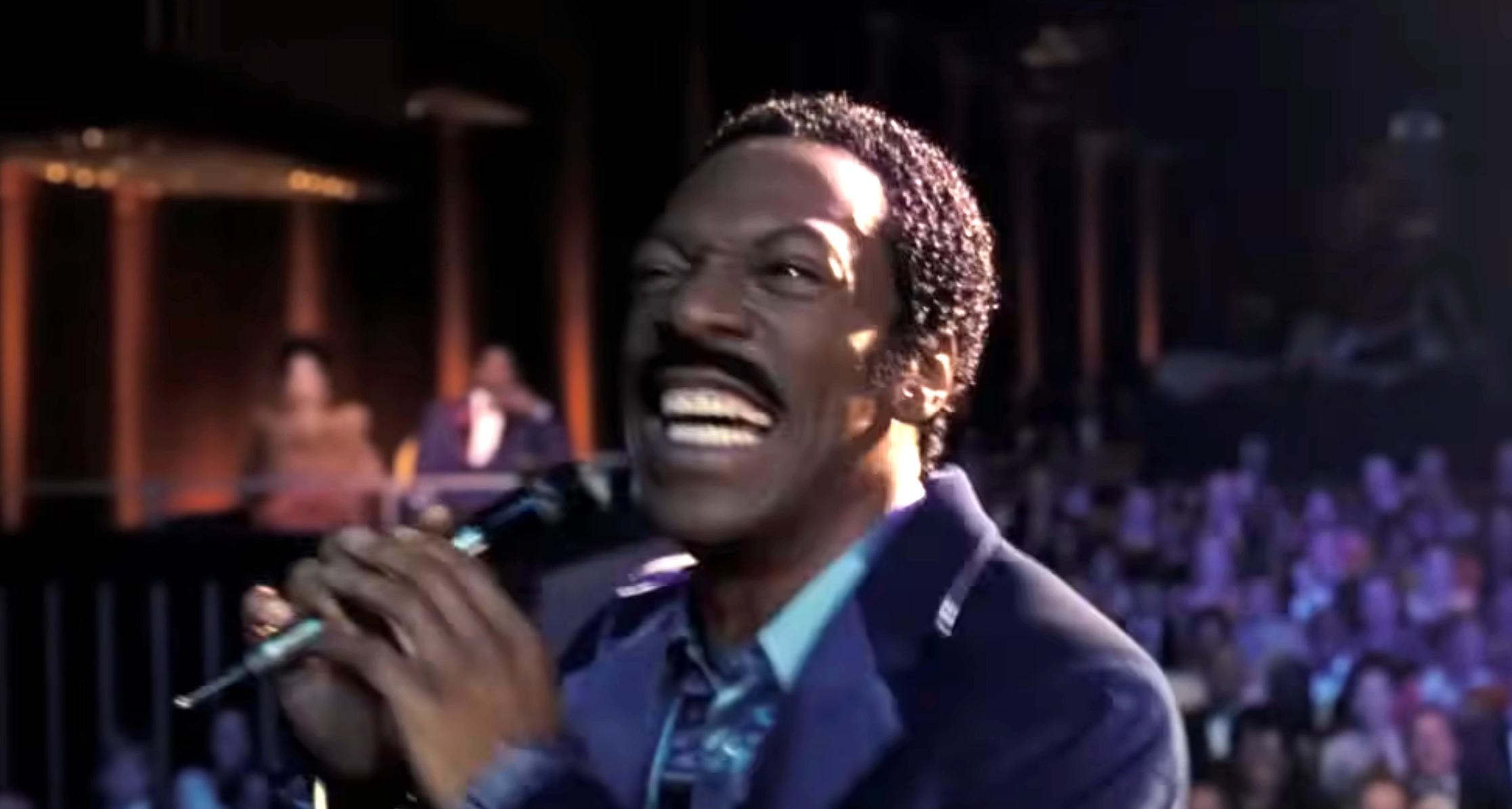 Eddie Murphy in a suit smiling and holding a microphone on stage, with an audience in the background