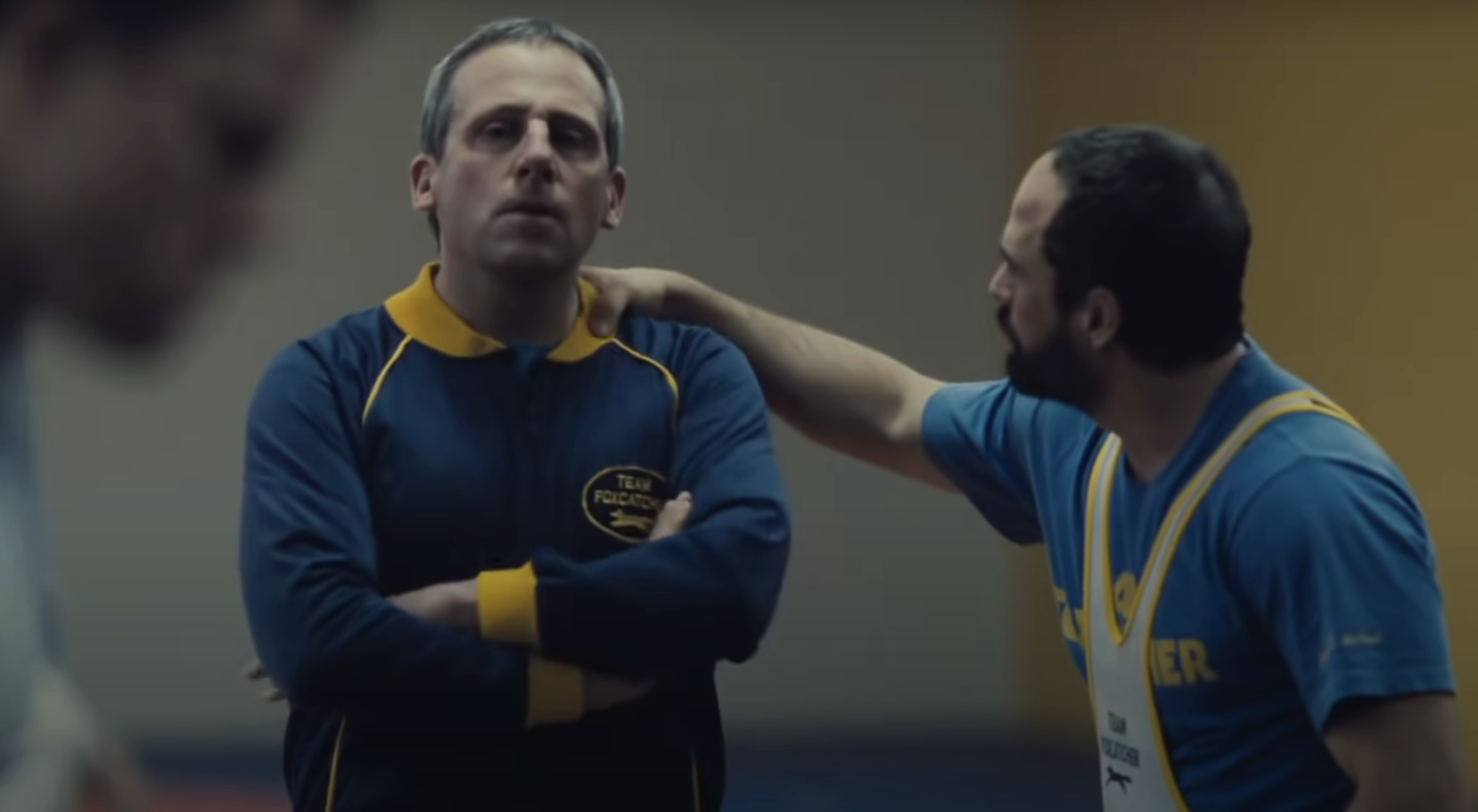 Steve Carell and Mark Ruffalo in workout clothes are standing in an indoor sports setting. The man is touching Carell&#x27;s shoulder, and Carell looks contemplative