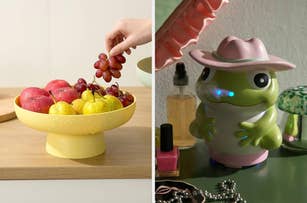 Hand placing grapes into a yellow fruit bowl beside a frog-shaped diffuser with a pink hat on a vanity table