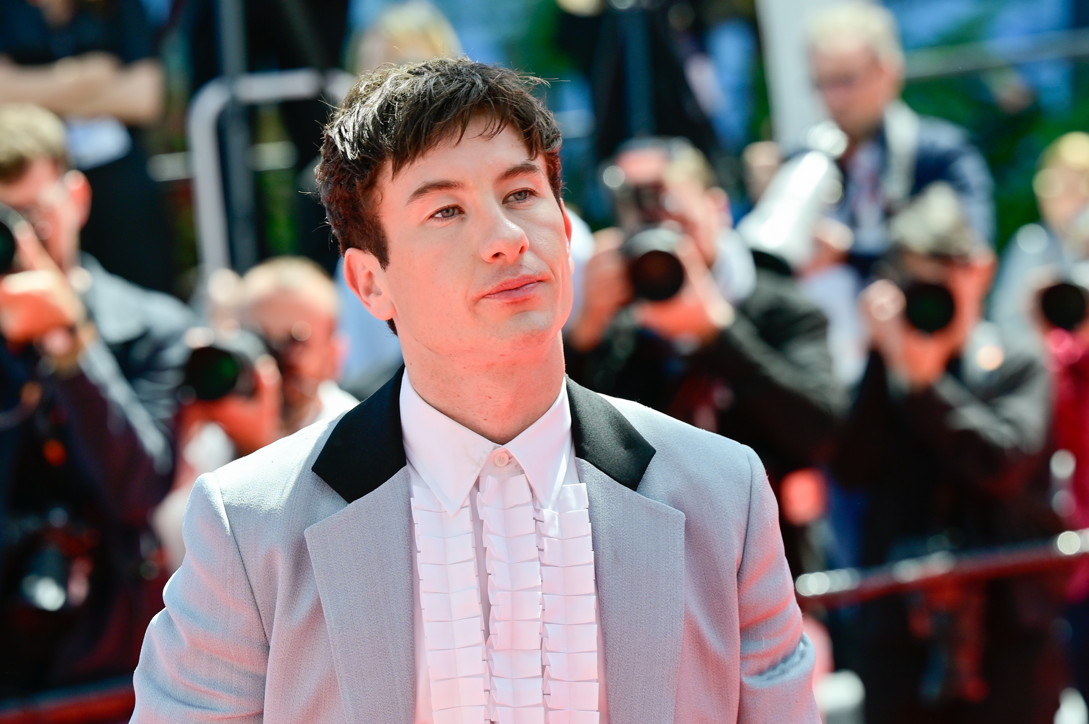 On the red carpet, Barry Keoghan is wearing a light-colored suit with a ruffled white shirt. Photographers are visible in the background