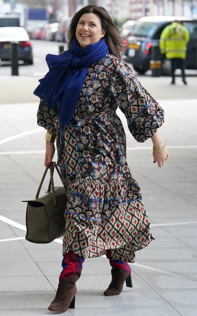 Person joyfully walking, wearing a patterned dress, blue scarf, and heeled boots, and carrying a brown handbag. Outdoor urban setting