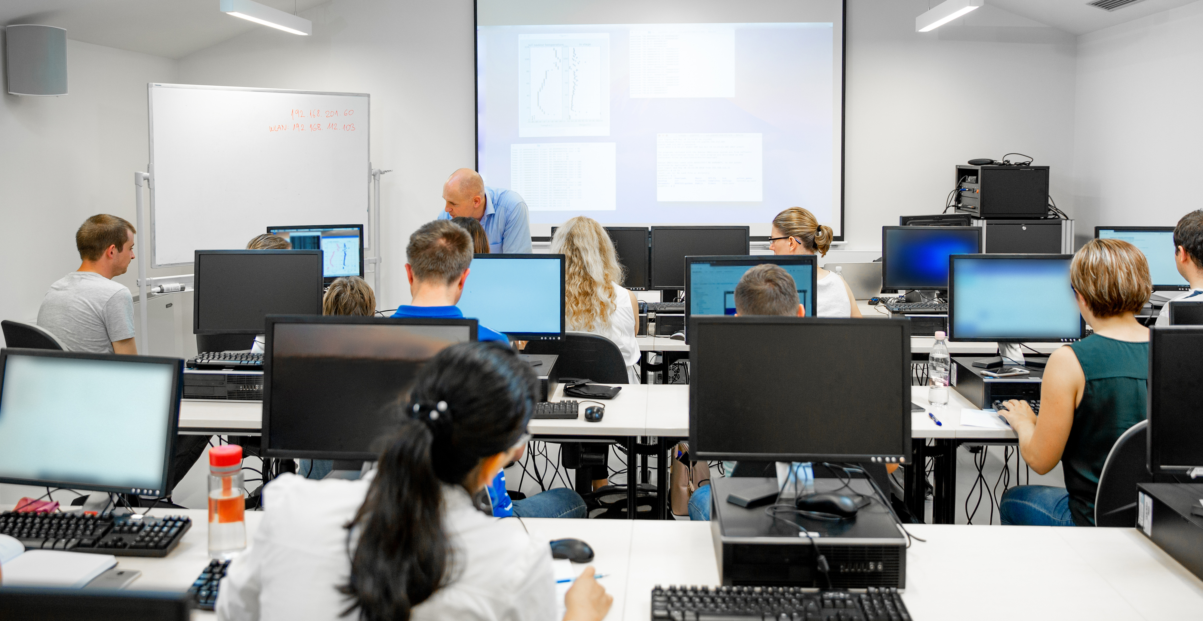 A group of people is seated in a training room at computer workstations, listening to an instructor at the front of the room