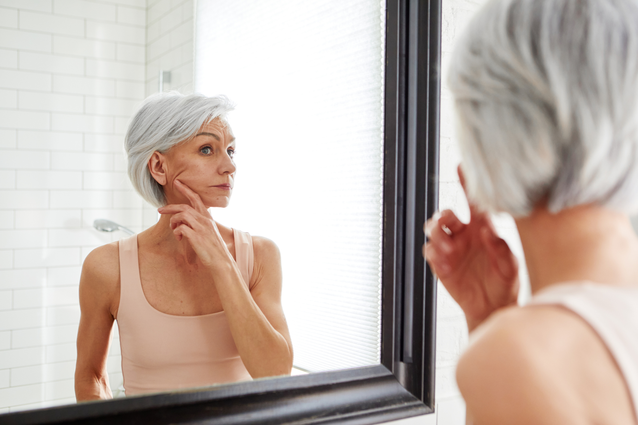 An older woman with short, gray hair is touching her face while looking into a bathroom mirror. She appears to be inspecting her skin