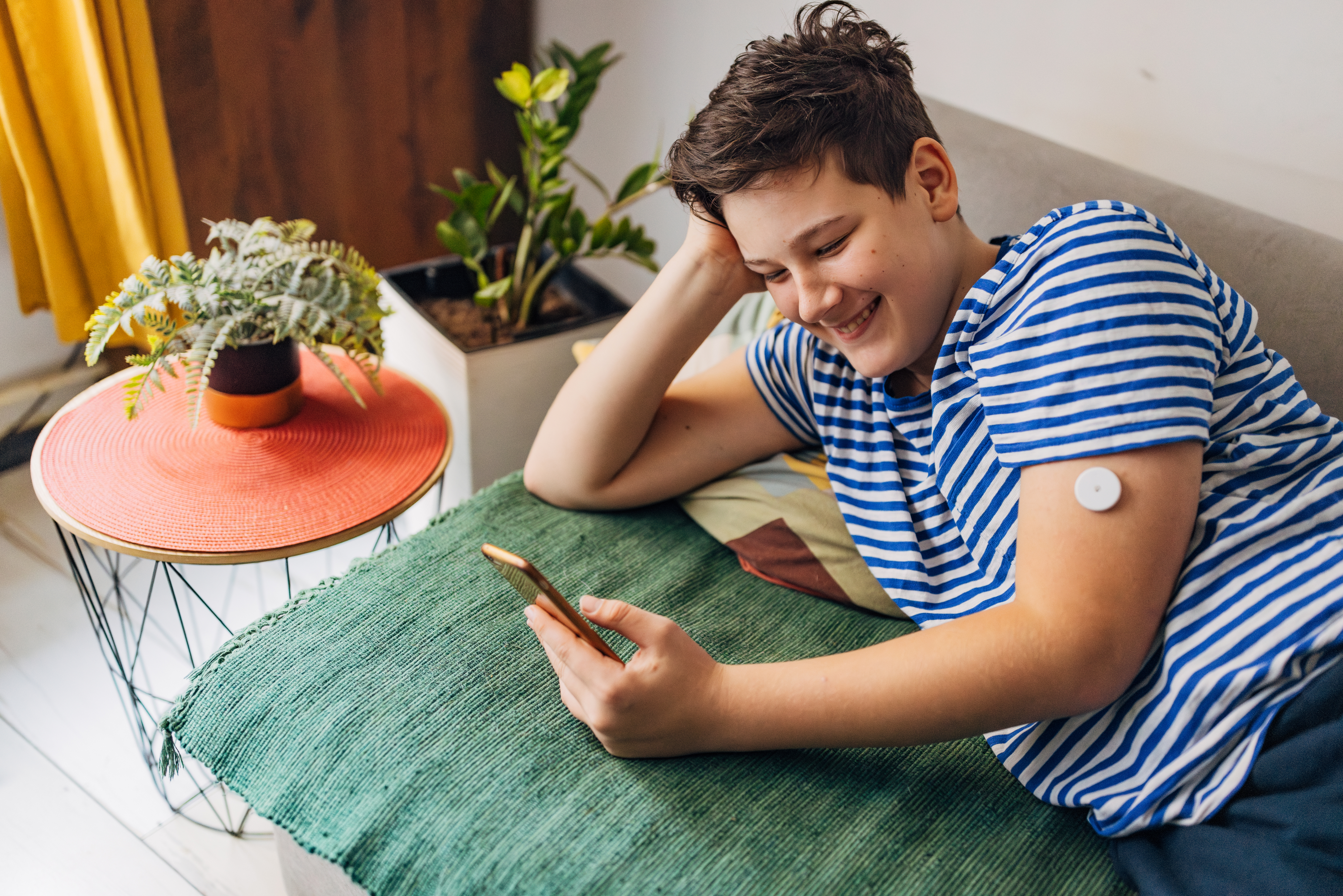 A person with a continuous glucose monitor on their arm is smiling while looking at their phone, relaxing on a couch with plants in the background