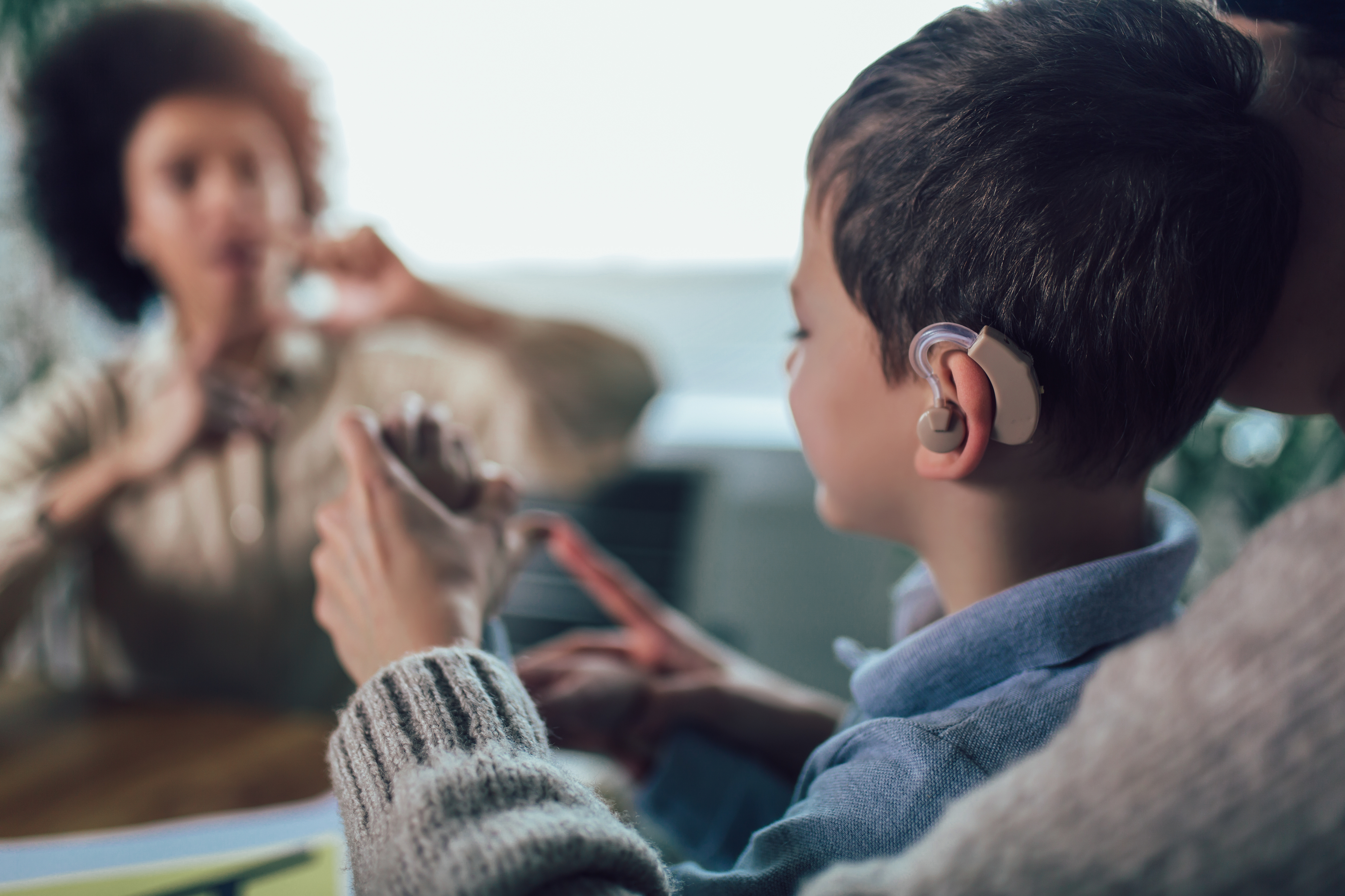 A young boy with a hearing aid communicates in sign language with a woman in the background, indicative of an inclusive and accessible learning environment