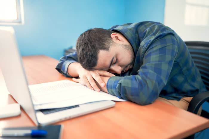 A person wearing a plaid shirt is asleep at a desk next to an open laptop and papers